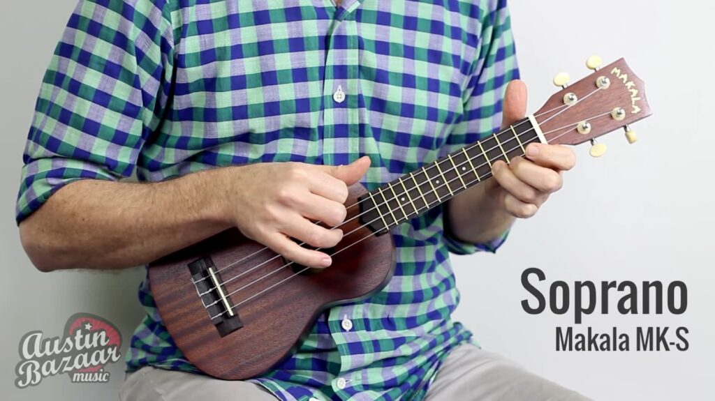 Close-up of hands playing a wooden Makala MK-S soprano ukulele, with a plaid shirt